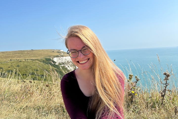 Photo of Jana Zeller smiling in front of a coastal landscape with cliffs and blue sea.