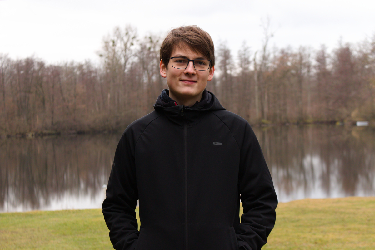 Photo of Jonas Teufel standing beside a calm lake with trees in the background.