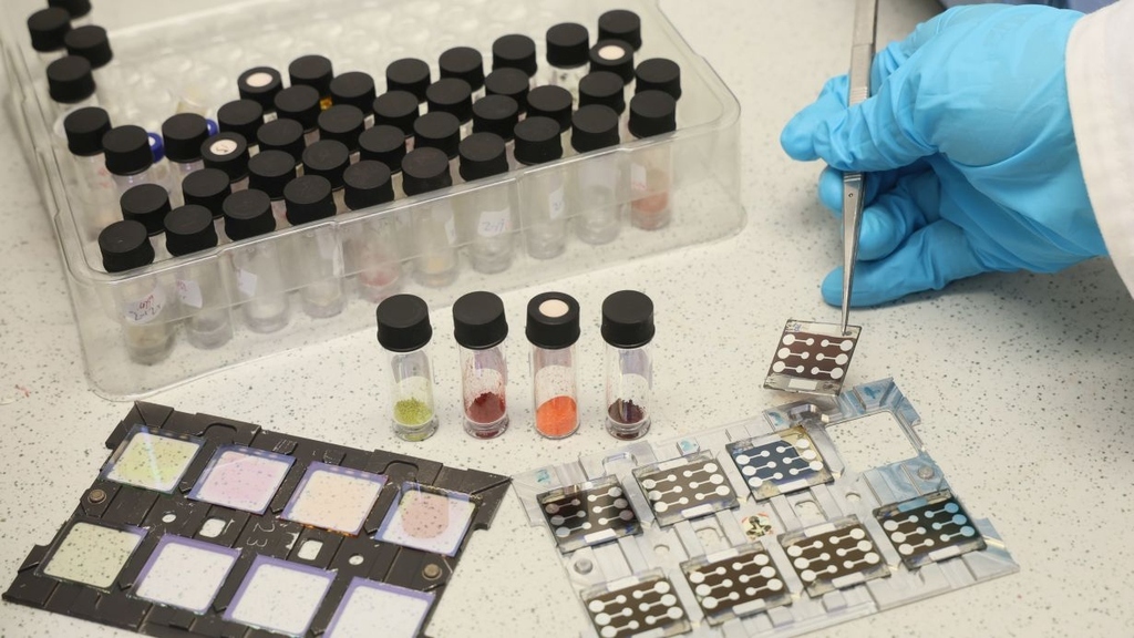 A gloved hand manipulates samples with tweezers on a lab bench with test tubes.